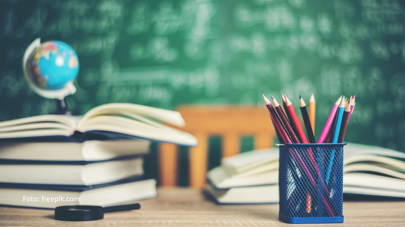 Ein Schreibtisch auf dem Bücher, Stifte und ein Globus steht. Im Hintergrund ist eine voll geschriebene Tafel.